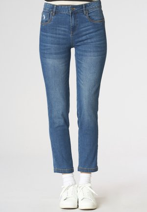 Jeans blu slim fit con tasche frontali, chiusura con bottone e cerniera, indossati con calzini bianchi alla caviglia e sneakers bianche.