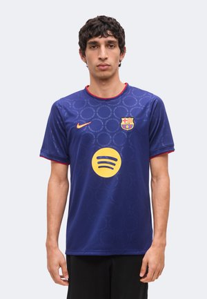 Jeune homme faisant face, portant un maillot de football bleu du FC Barcelone avec un logo Spotify jaune et un swoosh Nike orange.