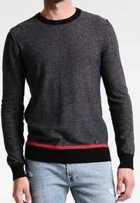 Grauer Strickpullover mit schwarzem geripptem Kragen und Bündchen, mit einem roten Streifen am Saum. Getragen mit verwaschenen hellblauen Jeans.