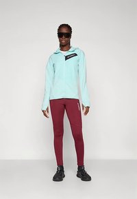 Kvinne iført lyseblå zip-up hettegenser, maroon leggings med "Terrex"-logo, svarte trening. sko og store svarte solbriller som står mot en ensfarget bakgrunn.