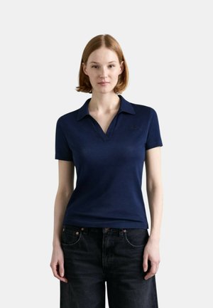 Giovane donna con capelli lisci fino alle spalle, che indossa una polo blu navy a maniche corte e jeans scuri, in piedi davanti a uno sfondo neutro.