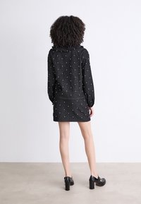 Tenue noire texturée comprenant un haut à manches longues avec un col à volants et une mini-jupe ajustée, ornée de détails en perles blanches.