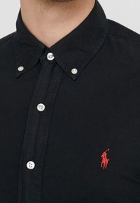 Muž v černé košili na knoflíky s malým červeným vyšitým logem polo hráče na hrudi.