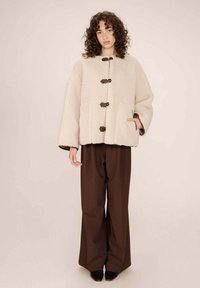Cappotto in shearling beige con orlo arrotondato, bottoni frontali e grandi tasche, abbinato a pantaloni larghi marroni e scarpe nere.
