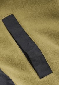 Fleecefabric i olivgrön med en svart texturerad remsa. Ytan är mjuk och visar en sydd kant och en framträdande, kontrasterande detalj.