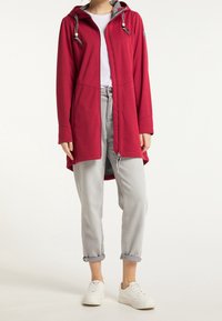 Personne portant un long sweat à capuche rouge zippé, un jean gris clair retroussé, des baskets blanches et un t-shirt blanc sur un fond uni.