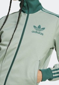 Grön zip-up-jacka med hög krage, med en mörkgrön adidas-logotyp, texturerat tyg och tre randiga detaljer på ärmarna.