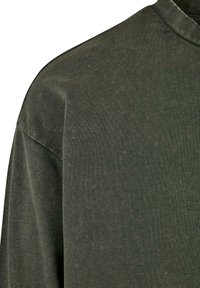 Tela de algodón verde oscuro mostrando el hombro y la manga superior de una camisa de manga larga con costura y puntadas visibles.