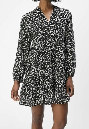 Femme portant une robe à manches longues à motif de feuilles noir et blanc, avec une jupe à volants et un col en V.