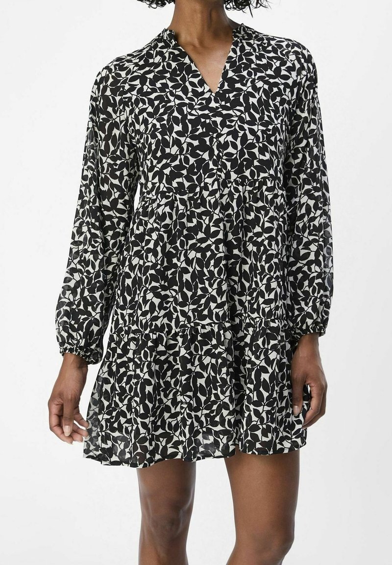 Femme portant une robe à manches longues à motif de feuilles noir et blanc, avec une jupe à volants et un col en V.