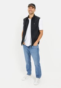 Czarna bezrękawnikowa kamizelka z przodem na zamek, noszona na białej koszulce, w połączeniu z niebieskimi dżinsami i białymi trampkami, na jednolitym tle.