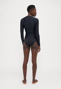 Dlhý rukáv čierny rash guard s priliehavým strihom, jemnou textúrou a kontrastným čiernym bikínovým spodkom. Pohľad zozadu zobrazuje elegantný siluetu.