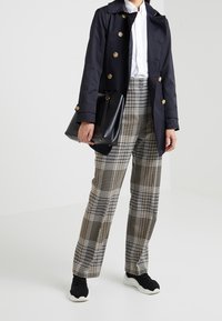 Osoba v námořnickém trenčkotu, bílém tričku, béžových a černých kostkovaných kalhotách, černých teniskách, držící černou kabelku na bílém pozadí.