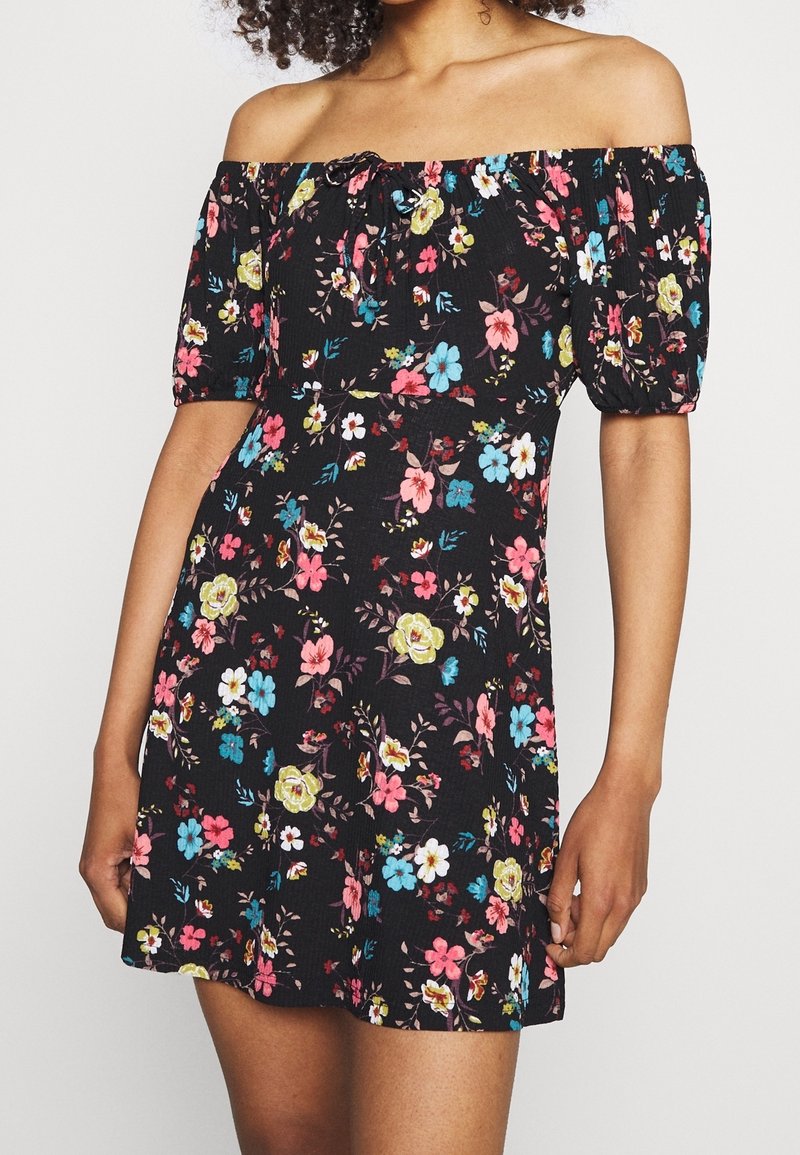 Mustassa kukkamekossa on pösöpuffihihat, olkapäät paljastava malli ja värikkäitä kukkakuvioita vaaleanpunaisena, sinisenä ja keltaisena.