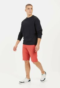 Marineblå sweatshirt med lange ærmer, parret med røde shorts. Model iført hvide sneakers med sorte accenter, stående foran en ensfarvet baggrund.