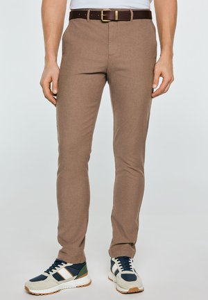 Mees, kes kannab kitsaste tumepruunide pükstega, tumedat pruuni nahkvööd, valget särki ja valgeid tossusid tumeroheliste ja siniste detailidega.