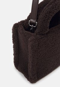 Bruine fleece handtas met een gestructureerde vorm, voorzien van leren hengsels en zilveren hardware. Zachte textuur met een gezellige uitstraling.