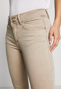 Beige farkkuhousut, joissa on keskimäinen vyötärö, kaksi etutaskua ja kontrastivärinen ompelu. Ominaisuudet kaksinkertainen napitus ja sileä pinta.