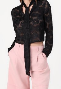 Černá průhledná blůza s květinovým vzorem, s kravatem u límce, zkrácený střih, doplněná vysokými růžovými kalhotami s širokými nohavicemi, hladká textura látky.