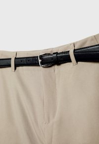Pantalon beige avec braguette zippée cousue et ceinture en cuir noir passée dans les passants avec une boucle argentée.