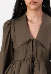 Robe vert olive avec un large col, détail de nœud sur le devant et taille froncée. Manches longues légèrement bouffantes. Tissu doux et texturé.