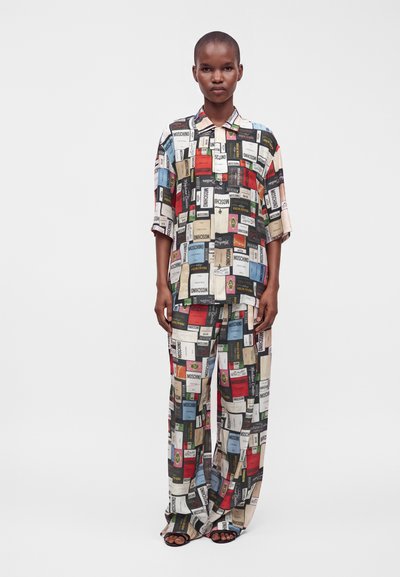 Modèle portant une chemise ample multicolore et un pantalon avec un imprimé graphique ressemblant à des étiquettes de produits éparpillées, se tenant devant un fond blanc uni.