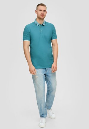Teal poloshirt met een gestreepte kraag en witte afwerking, gecombineerd met lichtblauwe distressed jeans en witte sneakers. Eenvoudige, casual look.