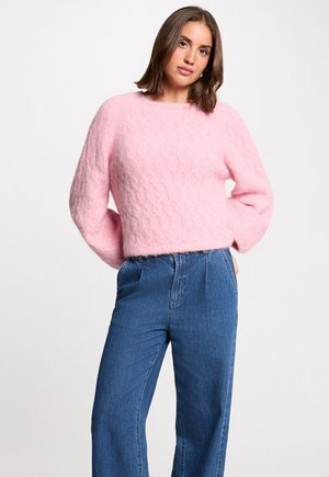 Kvinde iført en blød lyserød sweater og højtaljede blå jeans, stående med hænderne i lommerne mod en ensfarvet baggrund.