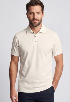 Man staat met één hand in zijn zak, draagt een beige polo met korte mouwen en een donkere broek tegen een effen achtergrond.