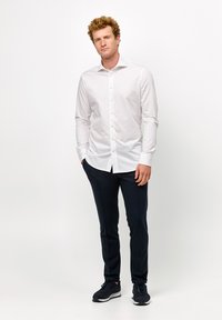 Weißes langärmliges Hemd mit Schalkragen, kombiniert mit marineblauen Hosen und dunklen Sneakers. Glatter Stoff mit maßgeschneiderter Passform.