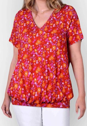 Vrouw draagt een rode V-hals blouse met oranje en roze bloemenprint en een witte broek, staand tegen een lichte achtergrond.