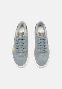 Reebok kedas ar pelēku zamšu augšdaļu, dzelteniem akcentiem, apaļu purngali, plakanu auklu un balto gumijas zoli. Perforēta purngala detaļa.