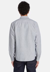 Camisa de manga larga gris claro con cuello doblado, puños abotonados y detalle de pliegue en la espalda. Tejido suave con una textura sutil.