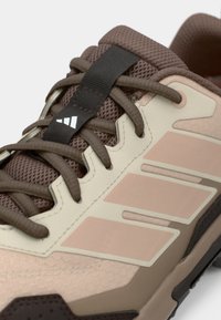 Αθλητικό παπούτσι ανοιχτό καφέ με υφή στην επιφάνεια, στοιχεία από δίχτυ και χαρακτηριστικές τρεις ρίγες σε απαλό ροζ. Διαθέτει καφέ κορδόνια και λογότυπο.