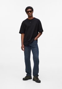 Volné černé tričko s malým logem, vzorované modré kalhoty s designem houndstooth a zelená obuv s tlustými podrážkami.
