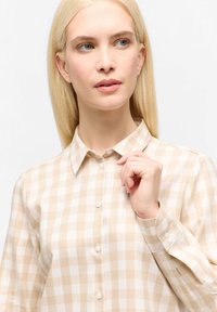 Blonde woman wearing beige and white checkered button-up shirt, holding collar with right hand, looking slightly right against white background.