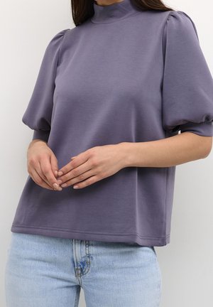 Frau trägt ein lila Puffärmel-Top mit hohem Kragen und hellblauen Jeans, die Hände zart vor dem Körper verschränkt.