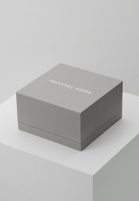 Scatola di cartone grigia con finitura testurizzata, con il logo bianco "MICHAEL KORS" sulla parte superiore, forma quadrata e design pulito e minimalista.