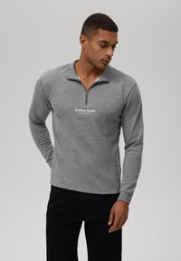 Sudadera gris con cremallera y mangas largas, hecha de una tela suave, cuenta con un logo blanco en el pecho y un pequeño cuello. Se lleva con pantalones oscuros.