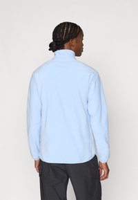 Hele sinine fliisjakk kõrge krae ja pikkade varrukatega, sileda tekstuuri ja mugava lõikega.