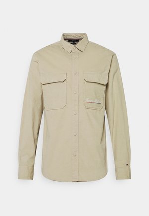 Beige long-sleeve button-up shirt with collar, two chest flap pockets, and subtle embroidered logo on the right pocket.