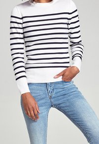 Persona che indossa un maglione bianco con strisce orizzontali nere e jeans azzurri chiari, con una mano in tasca, su uno sfondo semplice.