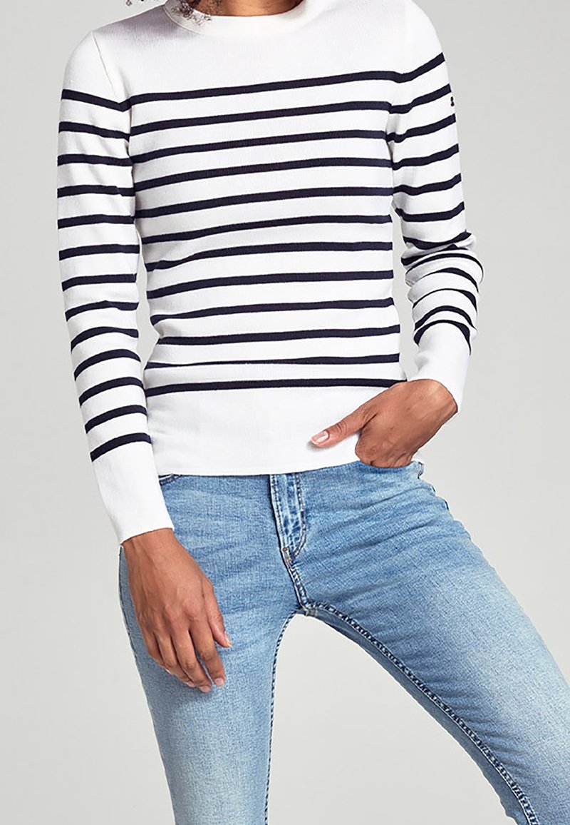 Persona che indossa un maglione bianco con strisce orizzontali nere e jeans azzurri chiari, con una mano in tasca, su uno sfondo semplice.