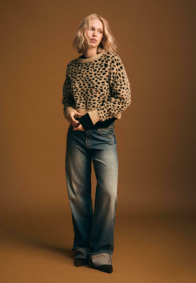 Beige Tierdruckpullover mit schwarzen Bündchen, kombiniert mit hellblauen weit geschnittenen Jeans und schwarzen High Heels. Weiche Textur, lockerer Schnitt.