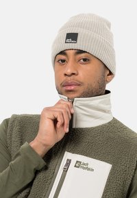 Beanie a coste grigio chiaro con logo, abbinato a un gilet in pile verde con zip caratterizzato da una tasca accentuata bianca e logo. Materiali texturizzati visibili.