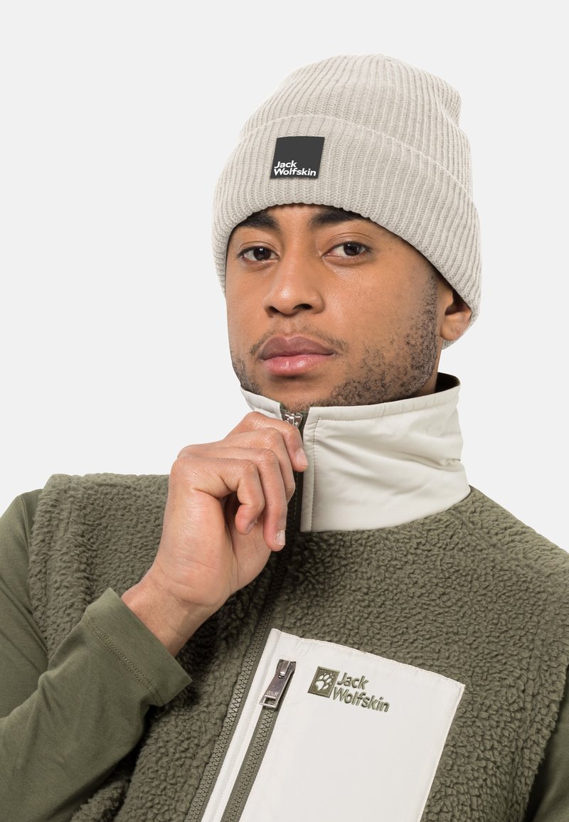 Beanie a coste grigio chiaro con logo, abbinato a un gilet in pile verde con zip caratterizzato da una tasca accentuata bianca e logo. Materiali texturizzati visibili.