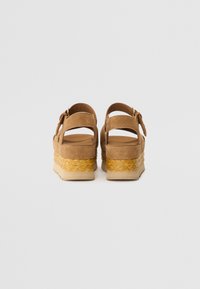 Sandalias de ante marrón con cuña trenzada al estilo espadrille y correa ajustable. Presenta un talón abierto y una plantilla en un marrón claro a juego.