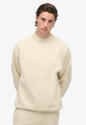 Jonge man met bruin haar, gekleed in een beige sweatshirt en bijpassende broek, staande met zijn handen achter zijn rug tegen een witte achtergrond.