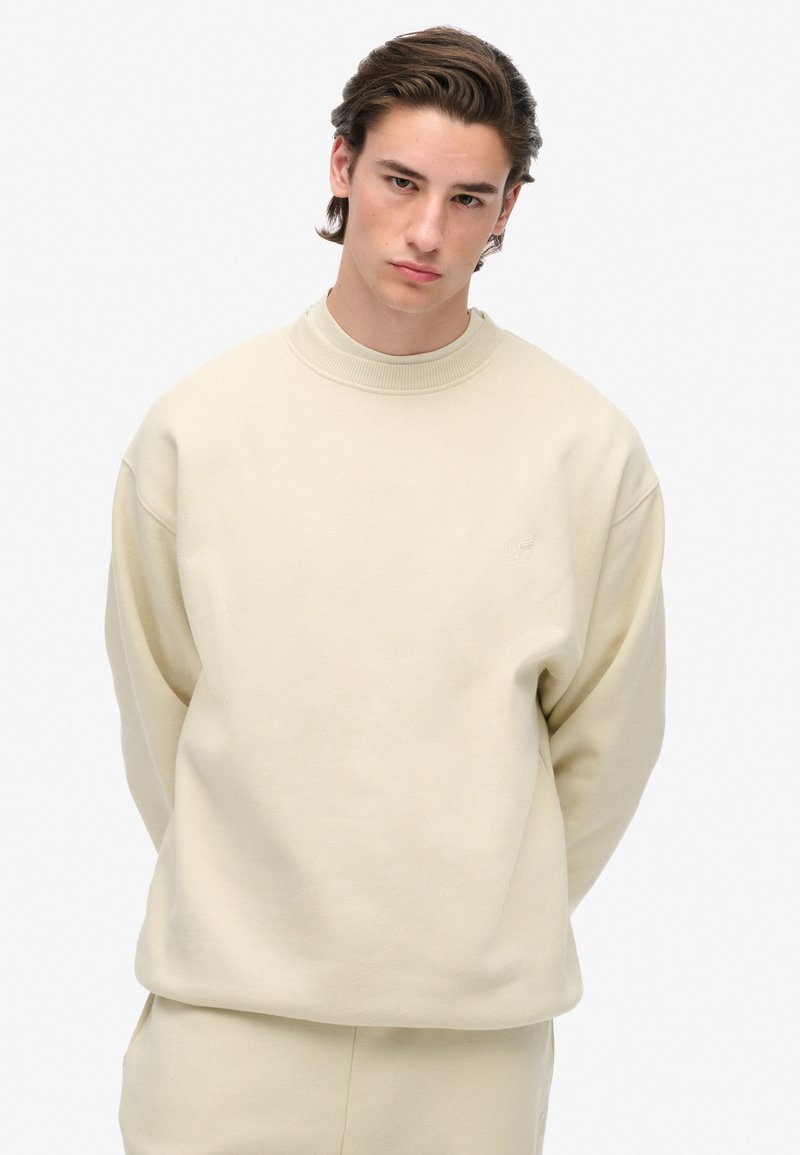 Giovane con capelli castani che indossa una felpa beige e pantaloni abbinati, in piedi con le mani dietro la schiena su uno sfondo bianco.
