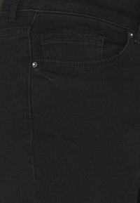 Jeans de ganga pretos com um primeiro plano do bolso de trás, com costura visível e um pequeno detalhe de rebite prateado. Textura suave.